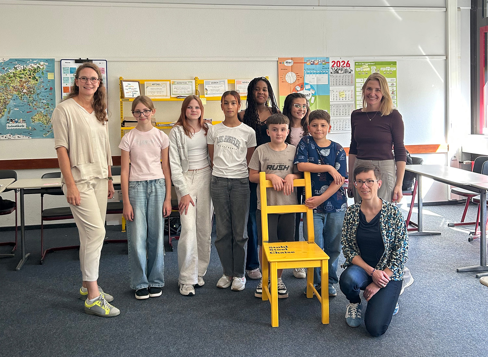 Lisa Wernsmann (Emsland-Gymnasium), die DaZ-Klasse des Emsland-Gymnasiums Rheine, Dr. Andrea Schneiker und Christina Çoban (beide VHS) bei der Stuhlübergabe am Emsland-Gymnasium Rheine (v.l.n.r.).